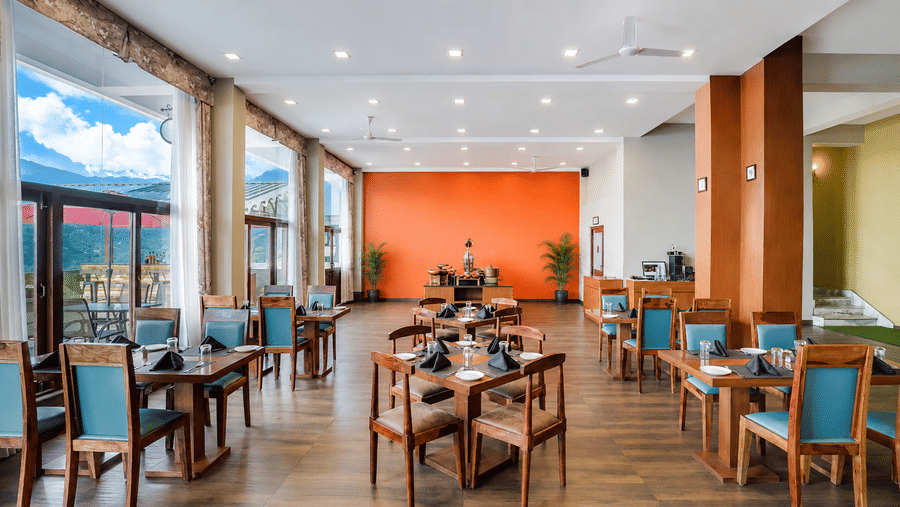 Restaurant at Valmere Himalayan Hotel & Spa, Gangtok with wooden dining tables, teal chairs, a buffet counter, an orange wall, and floor-to-ceiling windows.