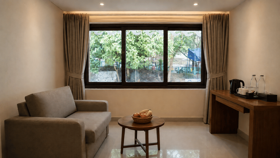 Sitting area at Valmere Himalayan Hotel & Spa, Gangtok with a gray sofa, a round wooden coffee table with a fruit bowl, and a desk with a kettle by the window.