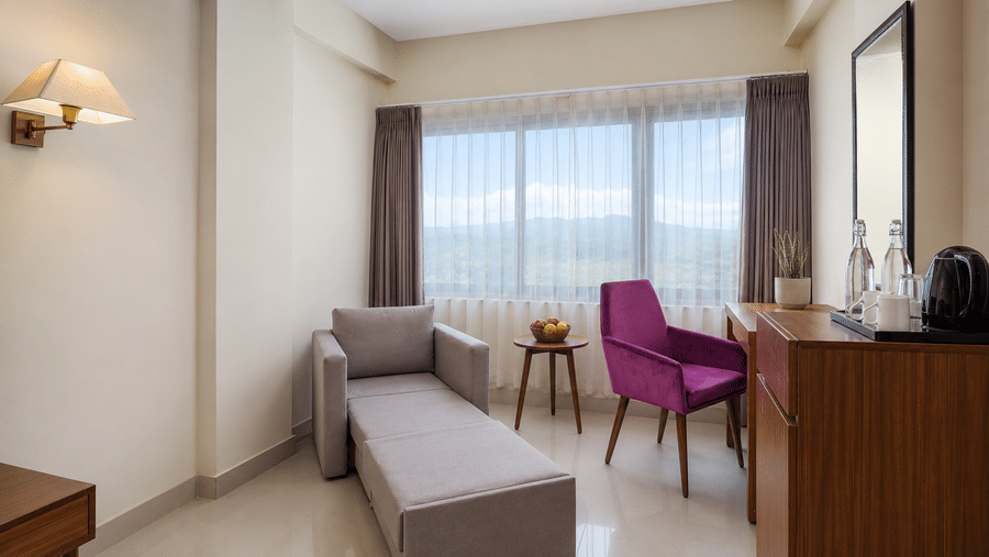 Sofa-cum-bed extended into a single bed at Valmere Himalayan Hotel & Spa, Gangtok, beside a purple armchair, round table, mirror, and a mountain-view window.