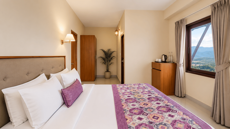 Suite bedroom at Valmere Himalayan Hotel & Spa, Gangtok with a king-size bed, purple bolster pillow, a wooden wardrobe, and a window framing a mountain valley view.