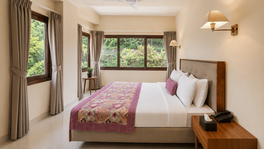 Suite bedroom at Valmere Himalayan Hotel & Spa, Gangtok with a king-size bed, purple runner, a nightstand telephone, and windows overlooking greenery.