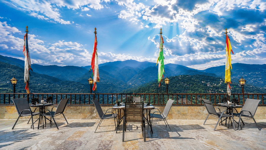 Terrace dining area at Valmere Himalayan Hotel & Spa, Gangtok with set tables, chairs, prayer flags, and a panoramic view of forested Himalayan mountains.