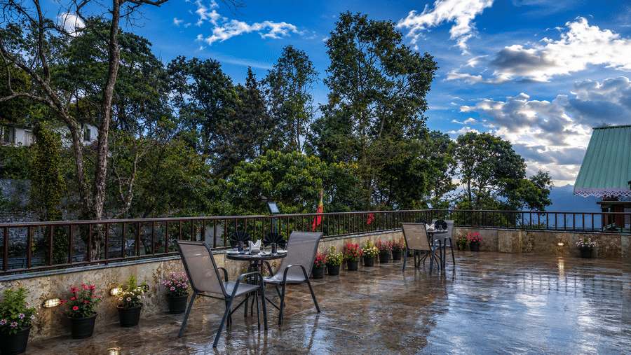 Terrace at Valmere Himalayan Hotel & Spa, Gangtok with 2 dining tables and chairs, potted flowering plants along the railing, and tall trees under a blue sky.