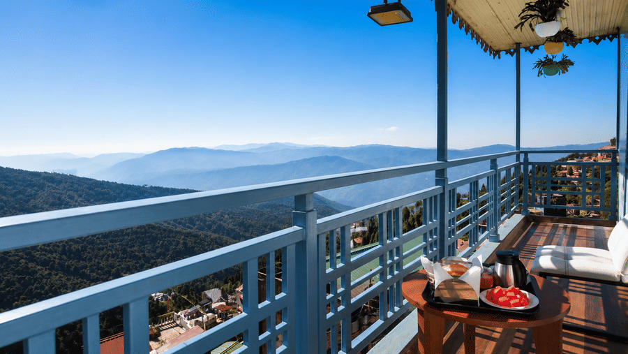 A wooden table with a food tray, a chair, and a balcony railing with panoramic views of mountains at Valmere Lungta Boutique Hotel & Spa, Darjeeling.