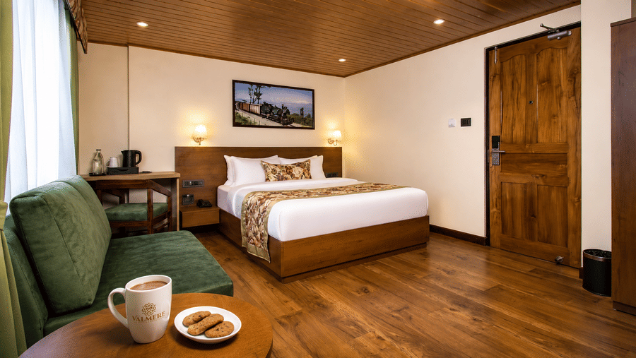 A king size bed, a sofa, a coffee table with a mug and biscuits, and a wooden door in a hotel bedroom at Valmere Lungta Boutique Hotel & Spa, Darjeeling.