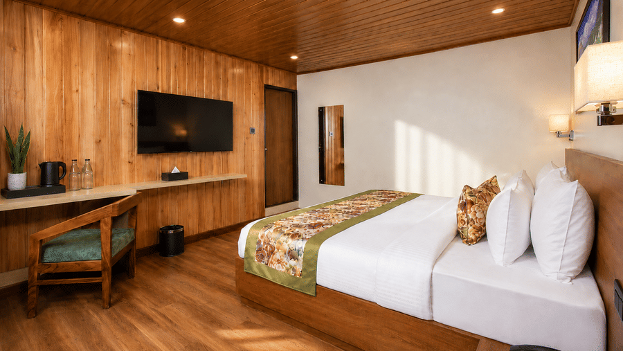 A king size bed, a wooden desk with a chair, a wall mounted television, and a mirror in a hotel bedroom at Valmere Lungta Boutique Hotel & Spa, Darjeeling.