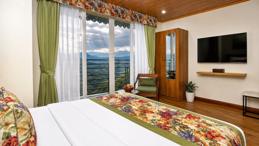 A king size bed, a television mounted on a wall, a wardrobe, and large glass windows with mountain views at Valmere Lungta Boutique Hotel & Spa, Darjeeling.