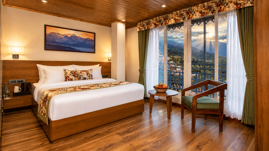 A king size bed, an armchair, a coffee table with fruits, and a window with mountain views in a hotel room at Valmere Lungta Boutique Hotel & Spa, Darjeeling.