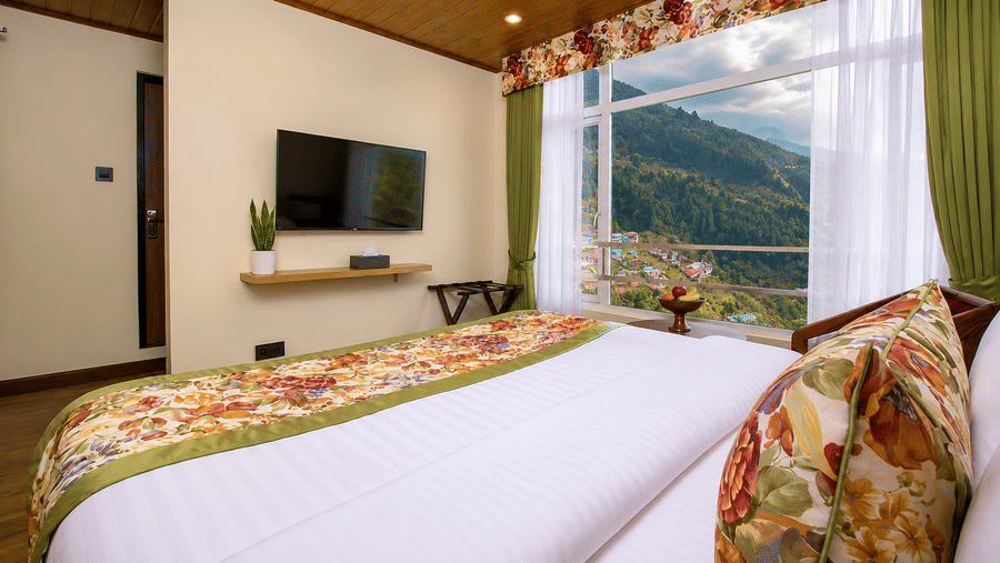 A large bed, a wall-mounted television, a wooden shelf, and a window with scenic valley views at Valmere Lungta Boutique Hotel & Spa, Darjeeling.