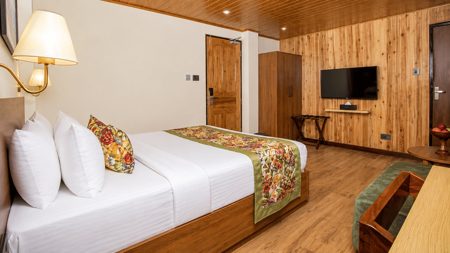 A king size bed, a wooden panelled wall with a television, a wardrobe, and a luggage rack in a hotel room at Valmere Lungta Boutique Hotel & Spa, Darjeeling.