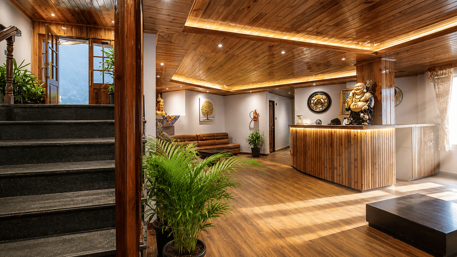 A wooden reception desk, a staircase, warm ceiling lights, and potted indoor plants at Valmere Lungta Boutique Hotel & Spa, Darjeeling.