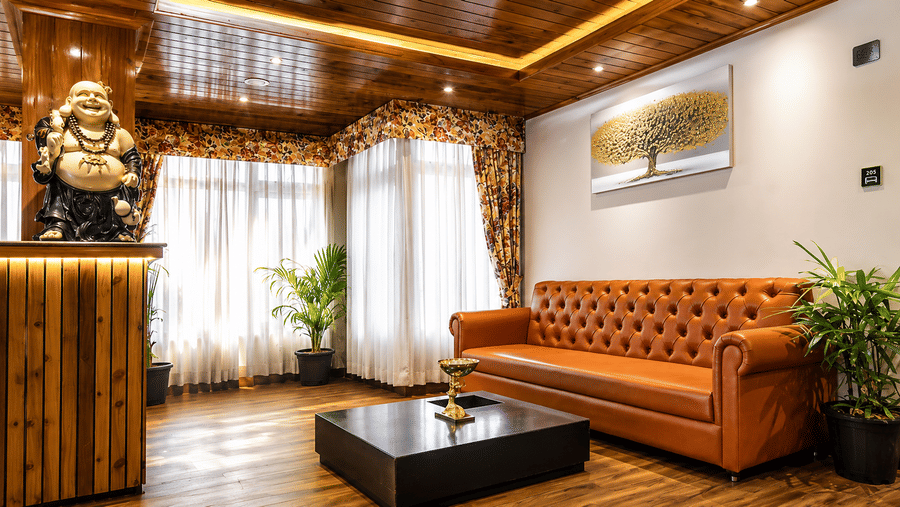 A tufted leather sofa, a wooden coffee table, a Buddha statue, and large curtained windows at Valmere Lungta Boutique Hotel & Spa, Darjeeling.