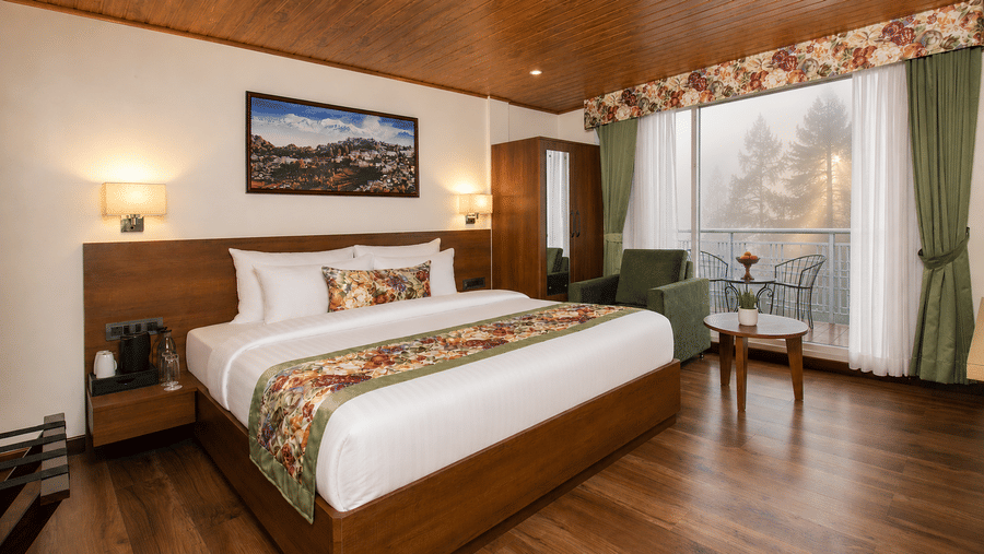 A king size bed, an armchair, a coffee table, and a wardrobe in a hotel bedroom with large windows at Valmere Lungta Boutique Hotel & Spa, Darjeeling.  