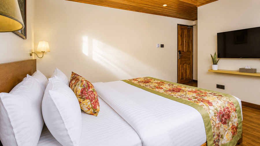 A king size bed with multiple white pillows, a floral bed runner, a wooden door, and a television at Valmere Lungta Boutique Hotel & Spa, Darjeeling.