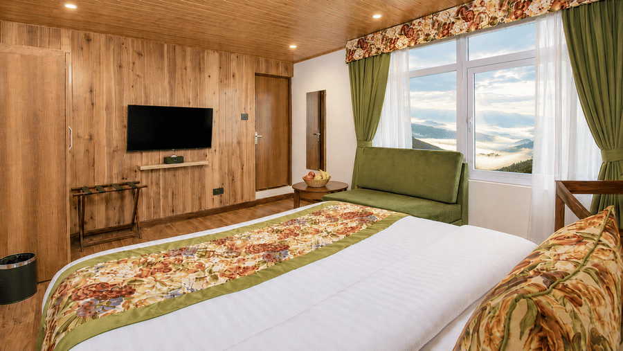 A double bed with floral bedding, a wall-mounted television, a sofa, and a large window showing mountain views at Valmere Lungta Boutique Hotel & Spa, Darjeeling.