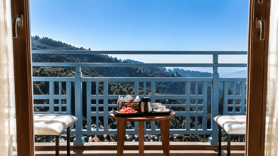 A round wooden table with a tea kettle, 2 chairs, and a metal railing on a balcony overlooking hills at Valmere Lungta Boutique Hotel & Spa, Darjeeling.