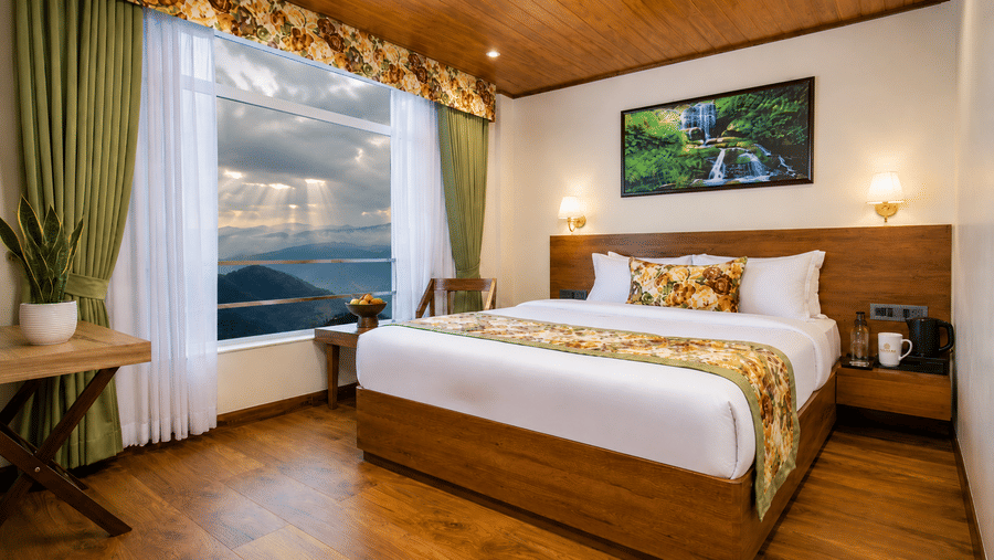 A king size bed, a wooden coffee table, a small potted plant, and a large window overlooking hills at Valmere Lungta Boutique Hotel & Spa, Darjeeling.