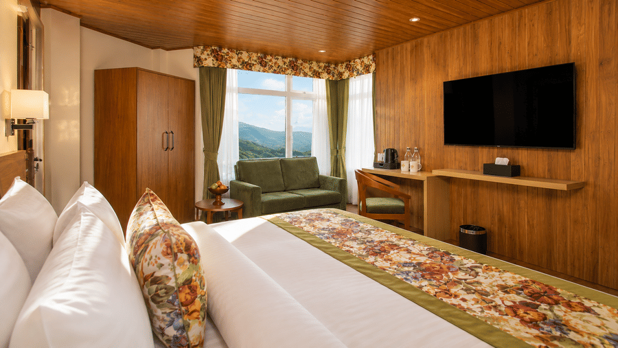 A king size bed, a sofa, a desk with a chair, and a wall mounted television in a hotel bedroom at Valmere Lungta Boutique Hotel & Spa, Darjeeling.