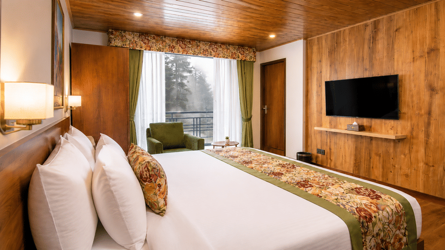 A king size bed, a wooden panelled wall, a television, an armchair, and a window at Valmere Lungta Boutique Hotel & Spa, Darjeeling.