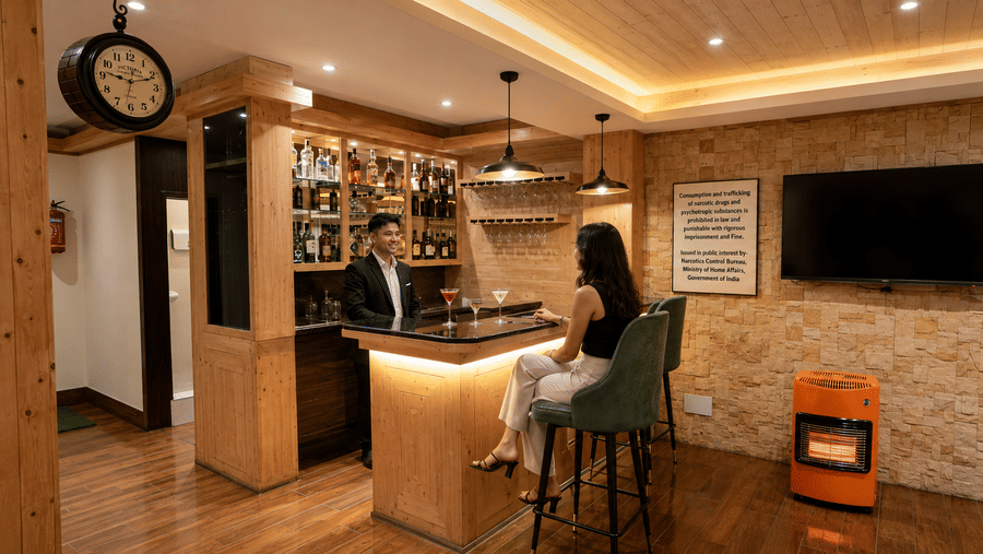 Bartender serving a guest at the bar counter at Valmere Magnolia Boutique Hotel & Spa, Pelling, with back lit liquor shelves, wine glasses, and a wall clock.