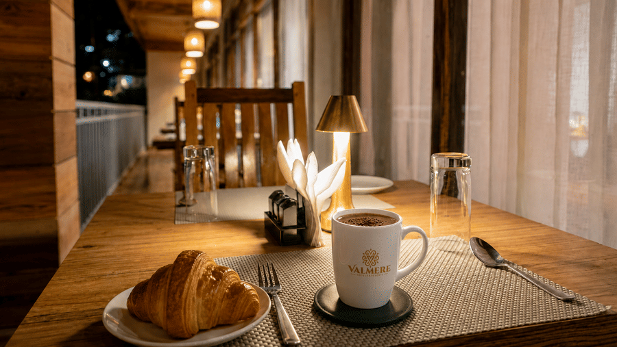 Branded coffee mug with hot chocolate and croissants on a table at Valmere Magnolia Boutique Hotel & Spa, Pelling, with a brass lamp and balcony view.