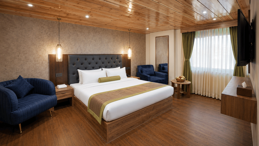 King Suite at Valmere Magnolia Boutique Hotel & Spa, Pelling with a wood-panelled bed, navy armchairs, a wall-mounted TV, and sheer curtains by the window.
