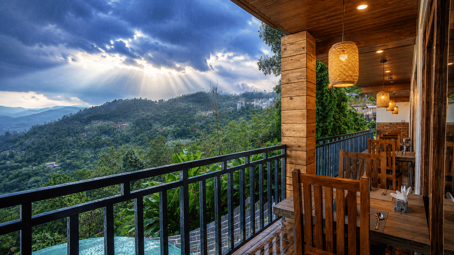 Outdoor dining balcony at Valmere Magnolia Boutique Hotel & Spa, Pelling with wooden furniture, rattan pendant lights, and a misty mountain valley view at dusk.