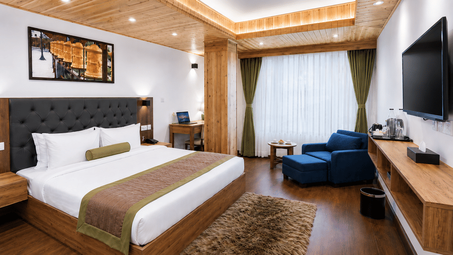 Guest room at Valmere Magnolia Boutique Hotel & Spa, Pelling with a bed, a navy armchair and ottoman by curtained windows, a wall-mounted TV, and a wooden desk.