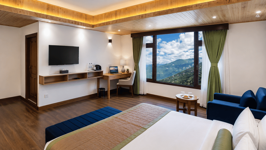 Guest room at Valmere Magnolia Boutique Hotel & Spa, Pelling with a bed, wooden desk, wall-mounted TV, navy armchair, and a window showing green mountains.