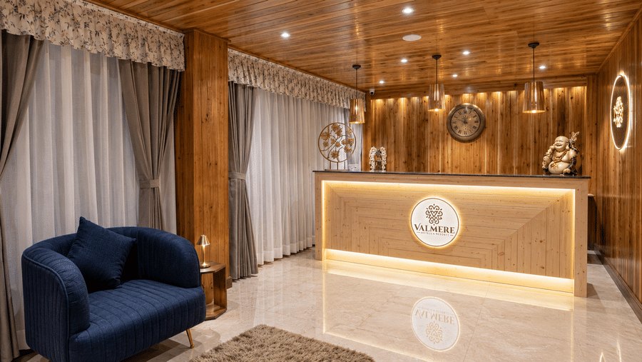 Reception desk at Valmere Magnolia Boutique Hotel & Spa, Pelling with a backlit wooden logo panel, wall clock, laughing Buddha statue, and an armchair.