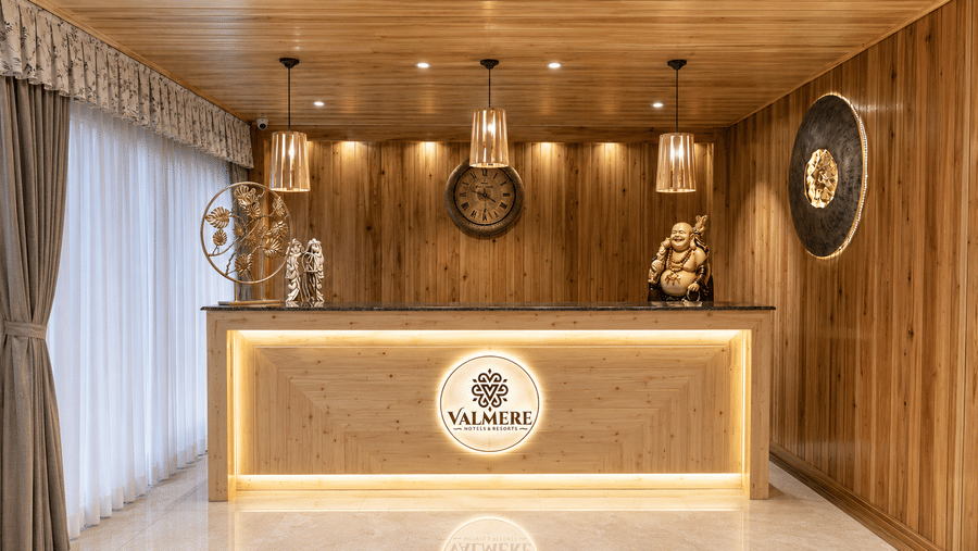 Straight-on view of the reception desk at Valmere Magnolia Boutique Hotel & Spa, Pelling with wood paneling, a wall clock, Buddha statue, and pendant lights.