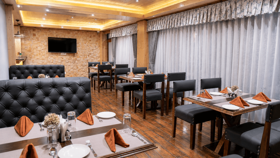 Restaurant seating at Valmere Magnolia Boutique Hotel & Spa, Pelling with black tufted booths, tables set with orange napkins, and a wall-mounted TV.