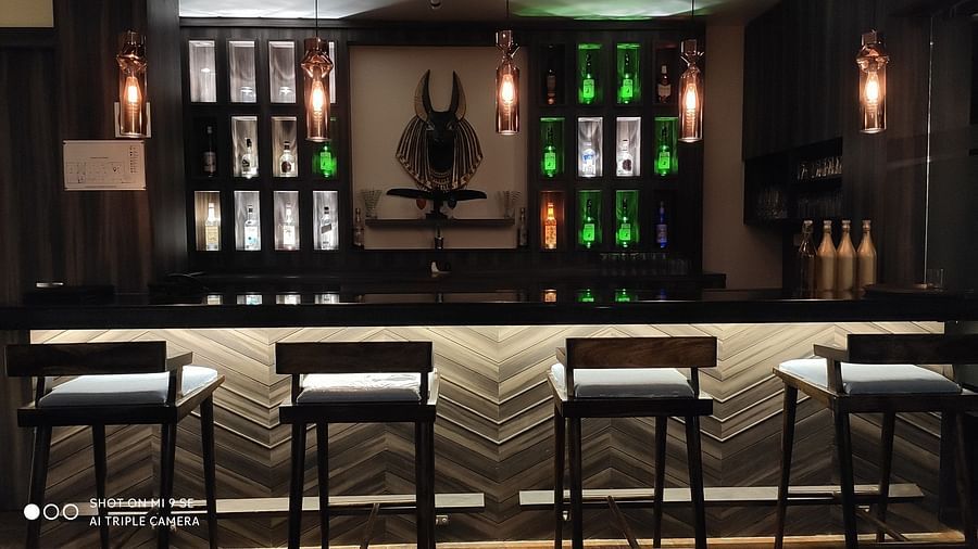 A modern bar area at night, featuring a dark counter, patterned bar stools, and back-lit shelves showcasing an array of bottles, some with a green glow - Sea Hills Hotels, Sri Vijaya Puram (Port Blair)