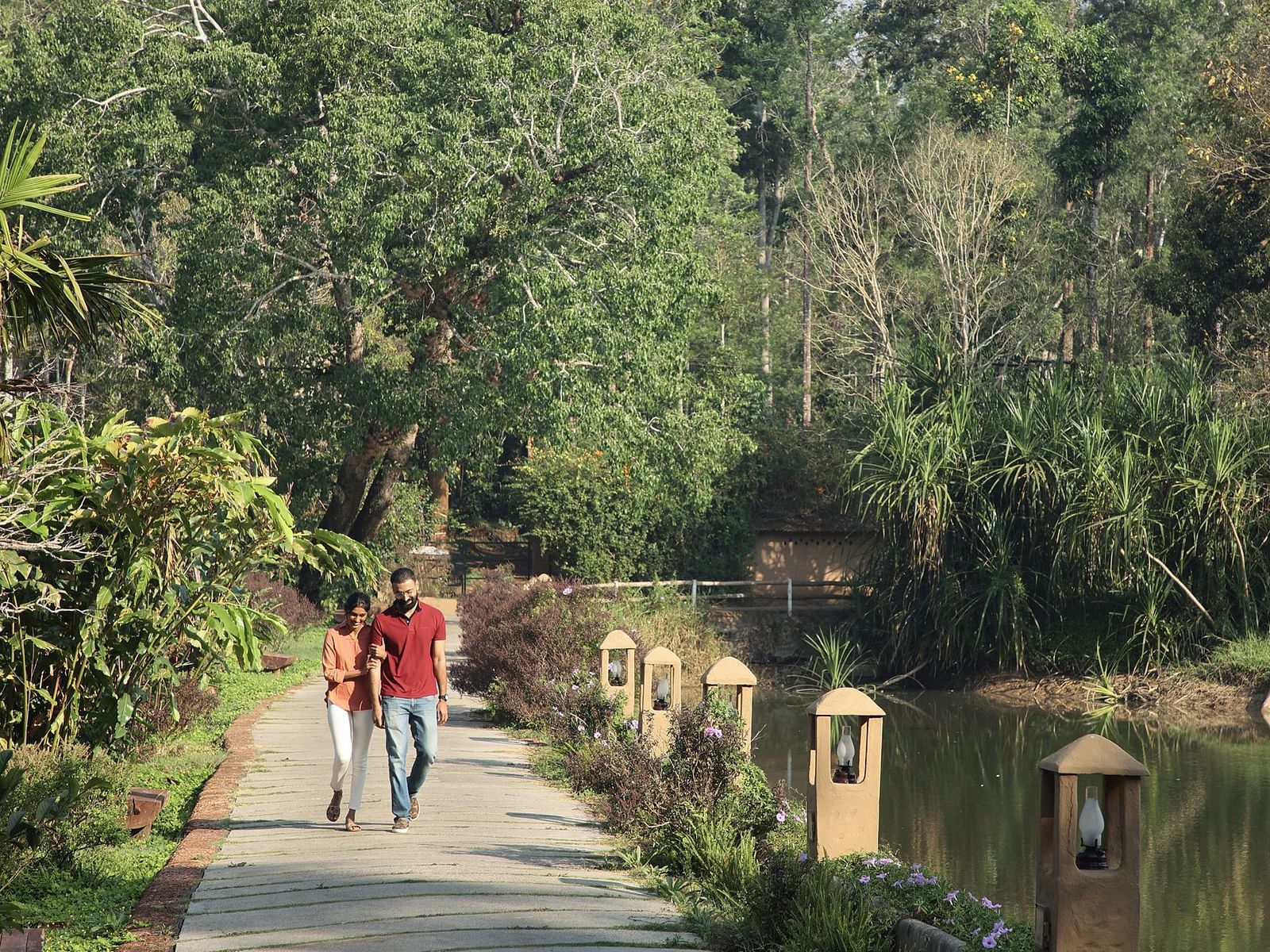 Two people walking on a path beside a pond in a lush, green park in Coorg