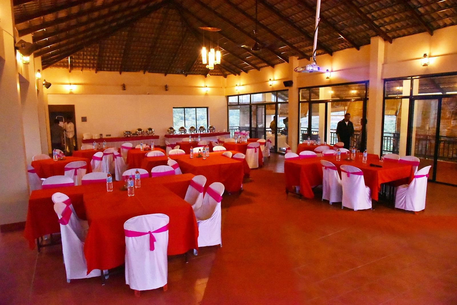 A banquet hall at Forest Escapes Koyna is prepared for a gathering with red tablecloths and white chair covers.