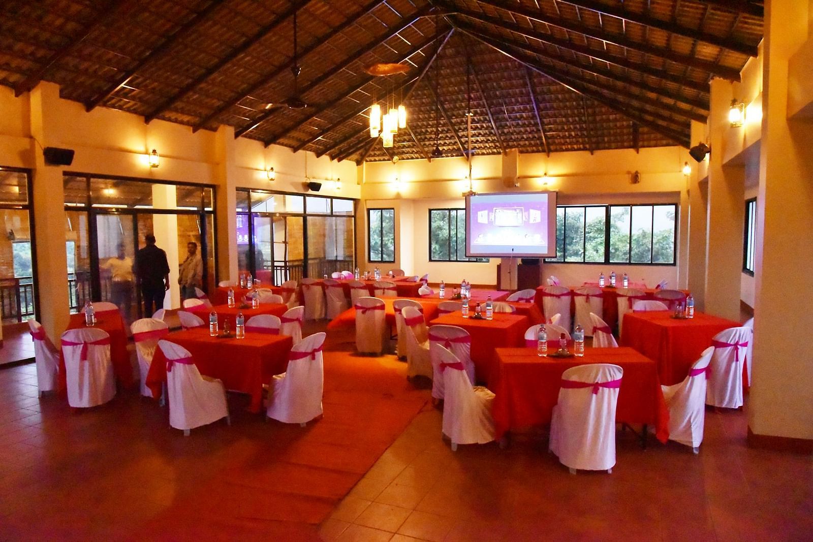 The spacious event hall at Forest Escapes Koyna is filled with round tables draped in red, prepared for a gathering.