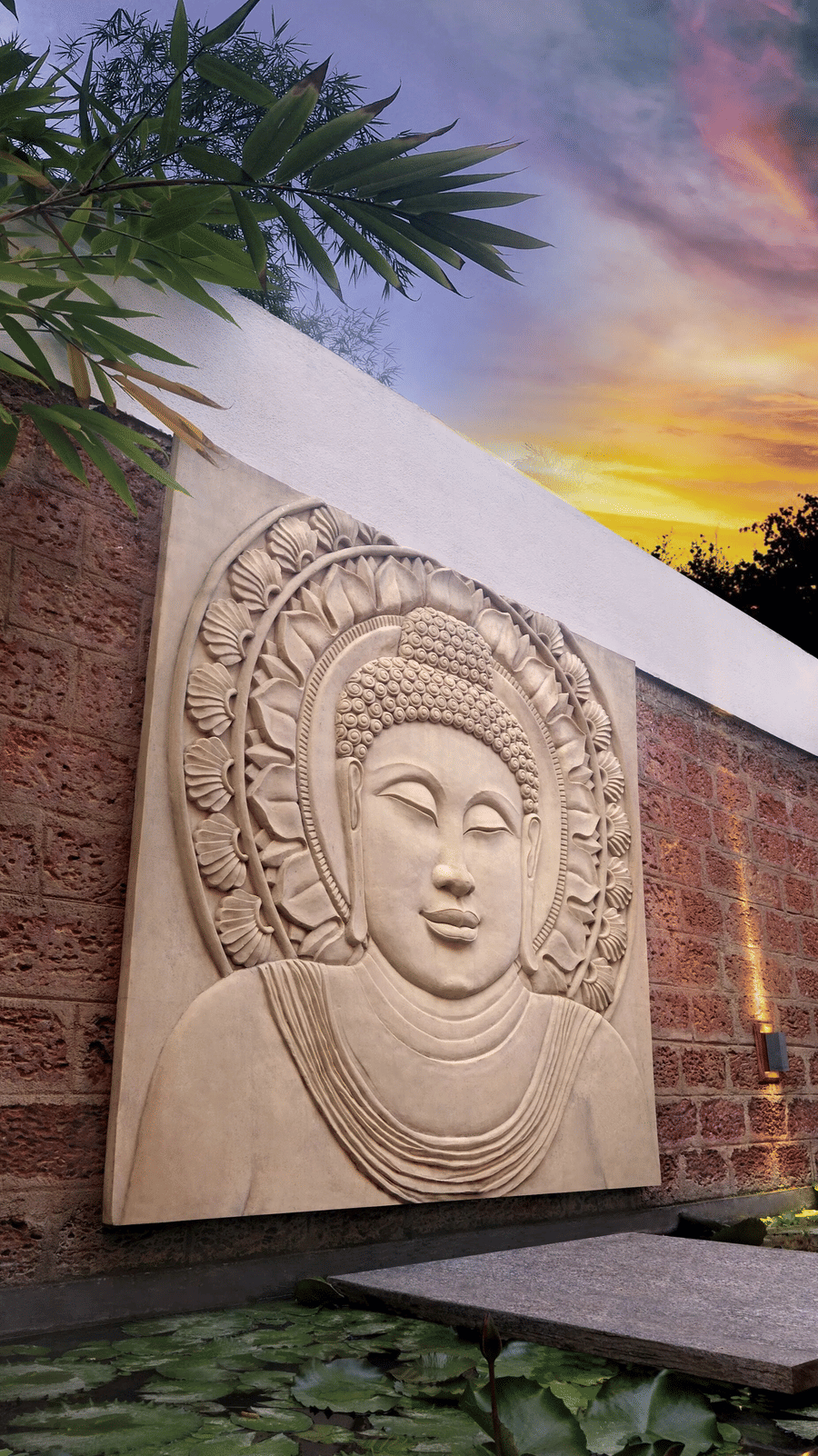 Stone Buddha carving on a textured wall by a lily pond under a twilight sky at Amanvana Spa Resort - A Luxury Resort in Coorg.