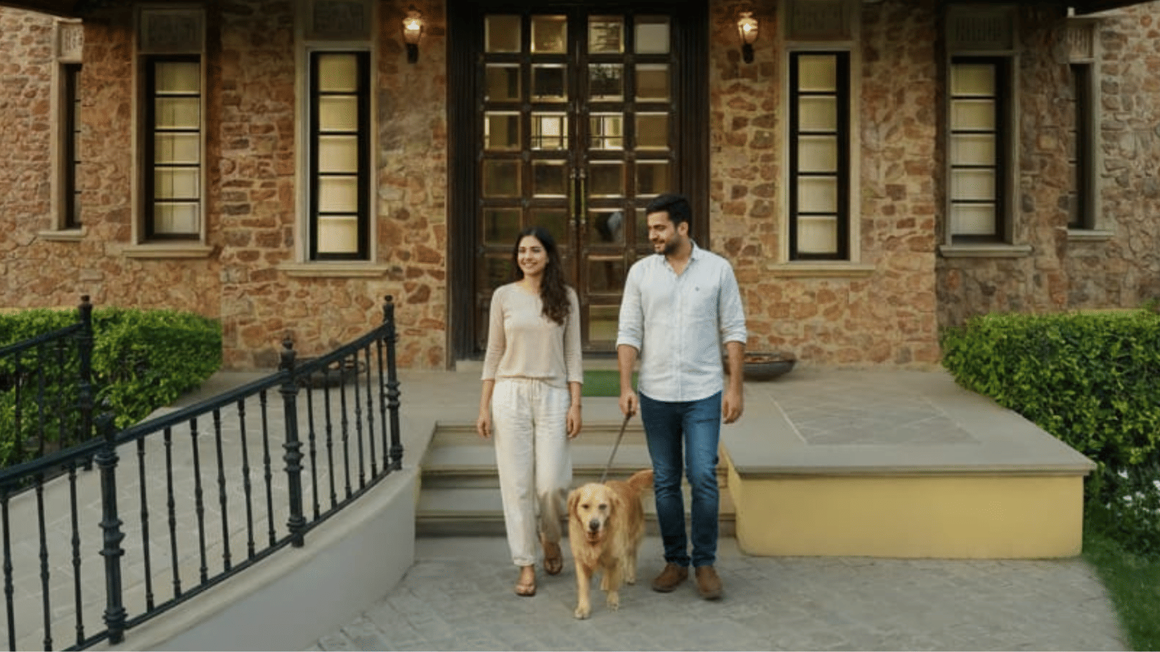 Young couple walking their golden retriever on the stone pathway outside Juna Mahal, with the heritage-style brick facade and manicured hedges in the background