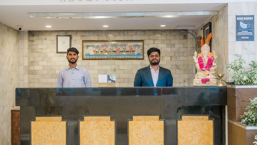 Two male staff members standing behind the front desk at Hotel Shiva Continental, Mussoorie, with a Ganesha idol, framed artwork, and signage in the background.