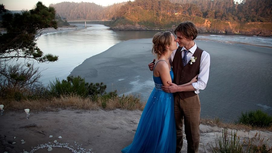 Bride in a blue gown and groom standing by a serene ocean at sunset