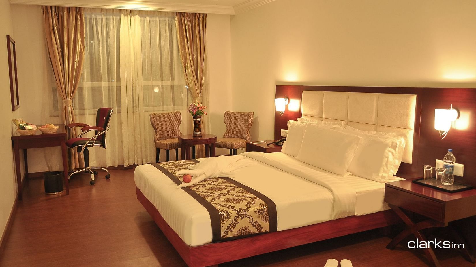 A warm-lit bedroom featuring a plush bed with nighstand and work desk as well as sitting space