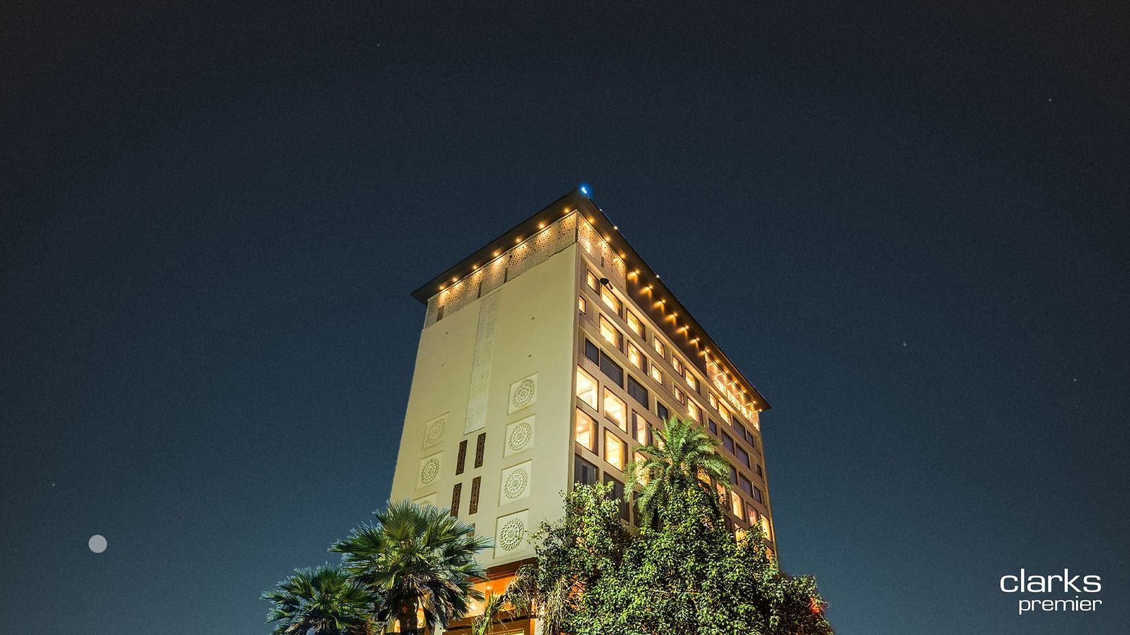 A shot of the top section of the facade of Clarks Premier, Kota in night sky lit up with warm lights and surrounded by trees