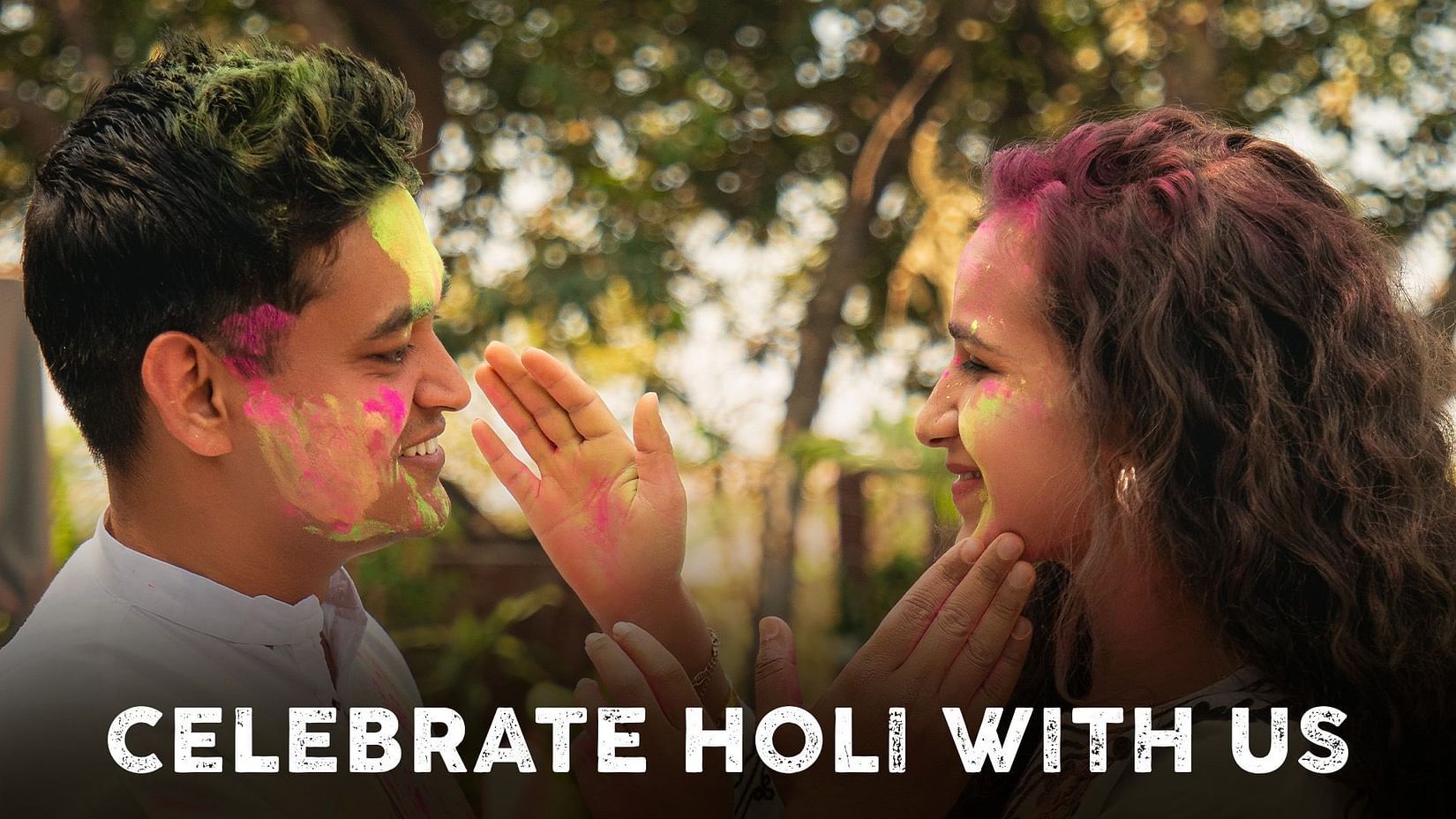 A man and a woman smile at each other, applying colorful powder to each others faces, with greenery in the background. Text reads Celebrate Holi with us.