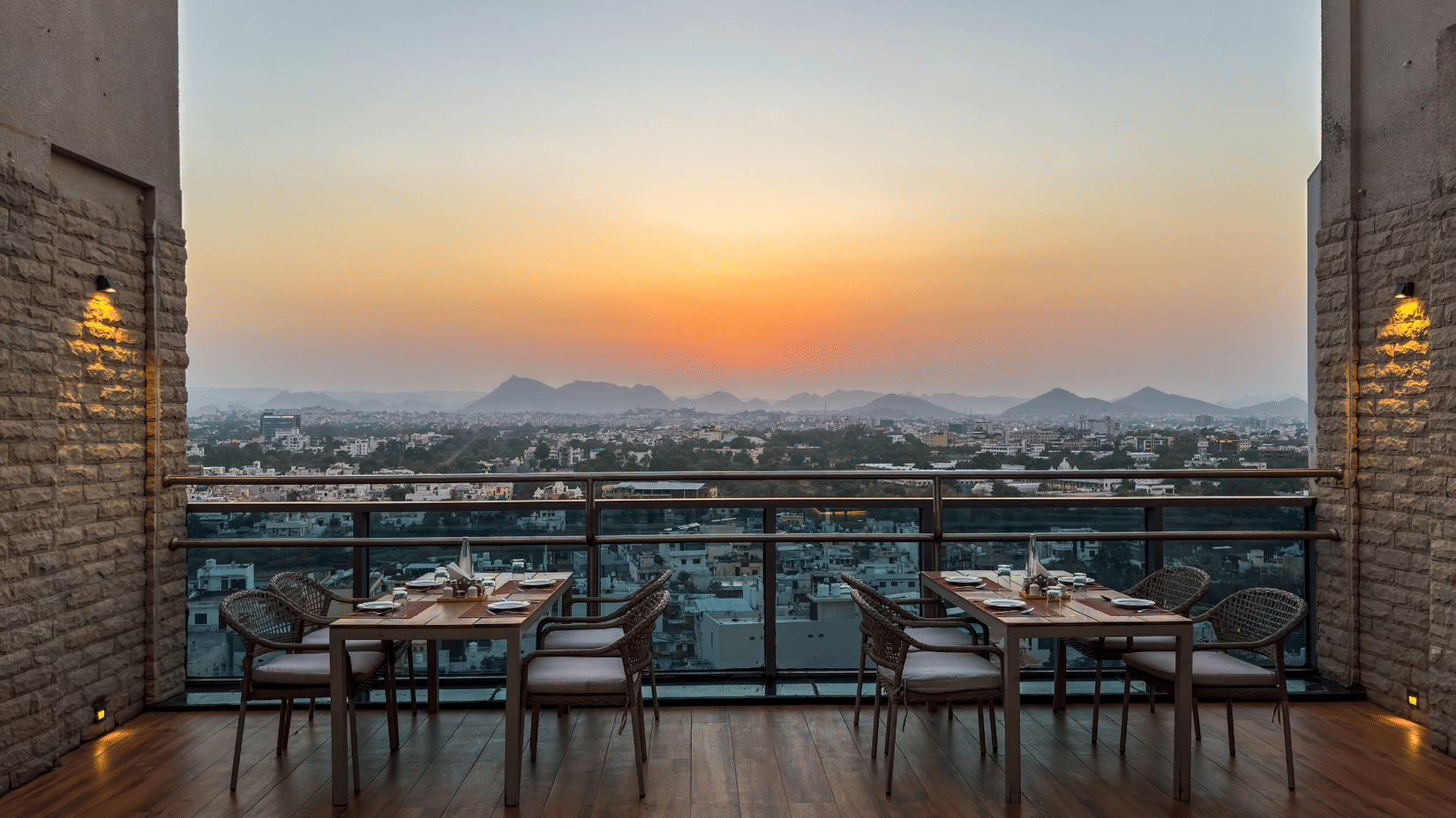 A view of a dining space at the Saltaire restaurant at Pride Premier Udaipur. Two setups of tables and chairs are positioned near a glass barrier overlooking a cityscape and distant hills.