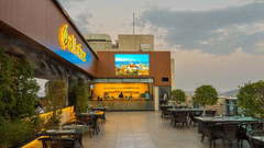 A view across the open terrace at the Saltaire restaurant at Pride Premier Udaipur. The area displays tables, chairs, potted plants, a bar counter towards the back, and a display screen mounted on a wall structure.