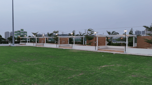 A wide outdoor green lawn space at Pride Resort, Rajkot featuring modern, structured shade elements and distant buildings under an overcast sky.
