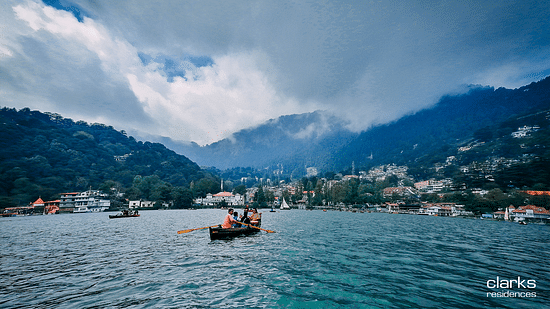 People boating on the Nainital lake amidst mountains and lush greenery under white and blue sky