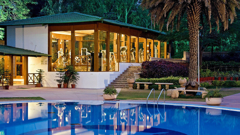 A glass-fronted fitness centre overlooking a swimming pool, surrounded by landscaped gardens and illuminated during the evening | Clarks Amer, Jaipur