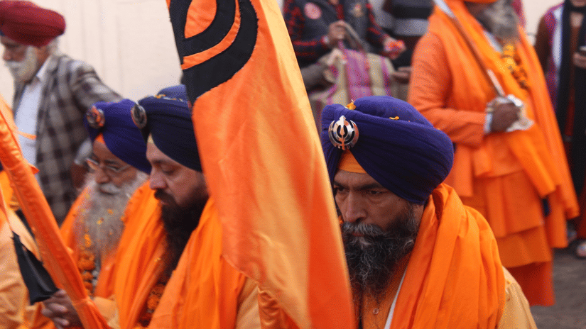 Sikh men in blue turbans and orange robes carrying Nishan Sahib flags during a religious procession or festival.