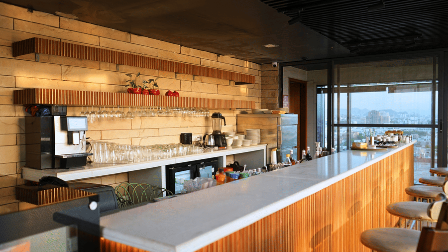 An interior view of a bar section within the Saltaire restaurant at Pride Premier Udaipur. The space contains a counter, bar stools, beverage preparation equipment, shelves on the wall, and windows looking out to the terrace.